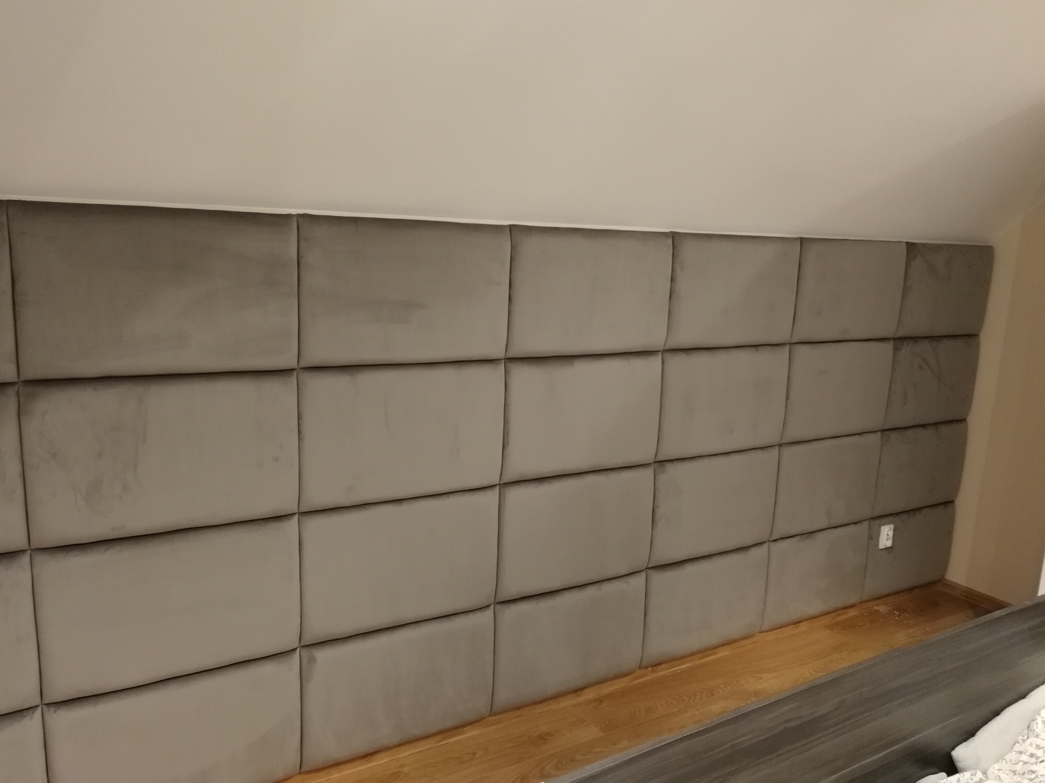 Panel tapicerowany w kolorze szarym, składający się z regularnych kwadratów, zamontowany na ścianie pod skosem, z widocznym gniazdkiem elektrycznym i fragmentem podłogi z paneli.