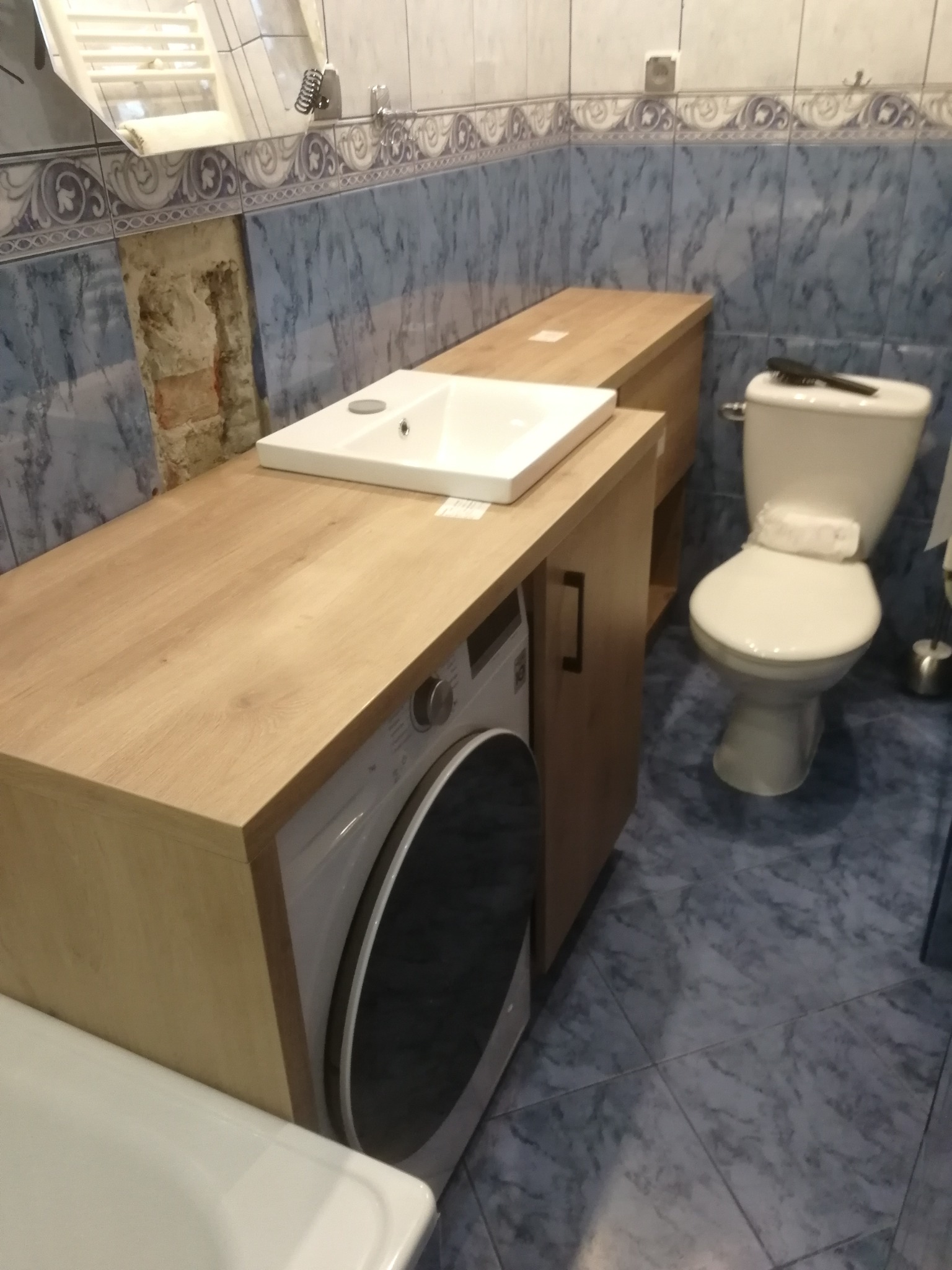 Łazienka w trakcie prac wykończeniowych: umywalka nablatowa na drewnianym blacie nad pralką, toaleta z deską, niebieskie płytki imitujące marmur, częściowo odsłonięta cegła na ścianie.