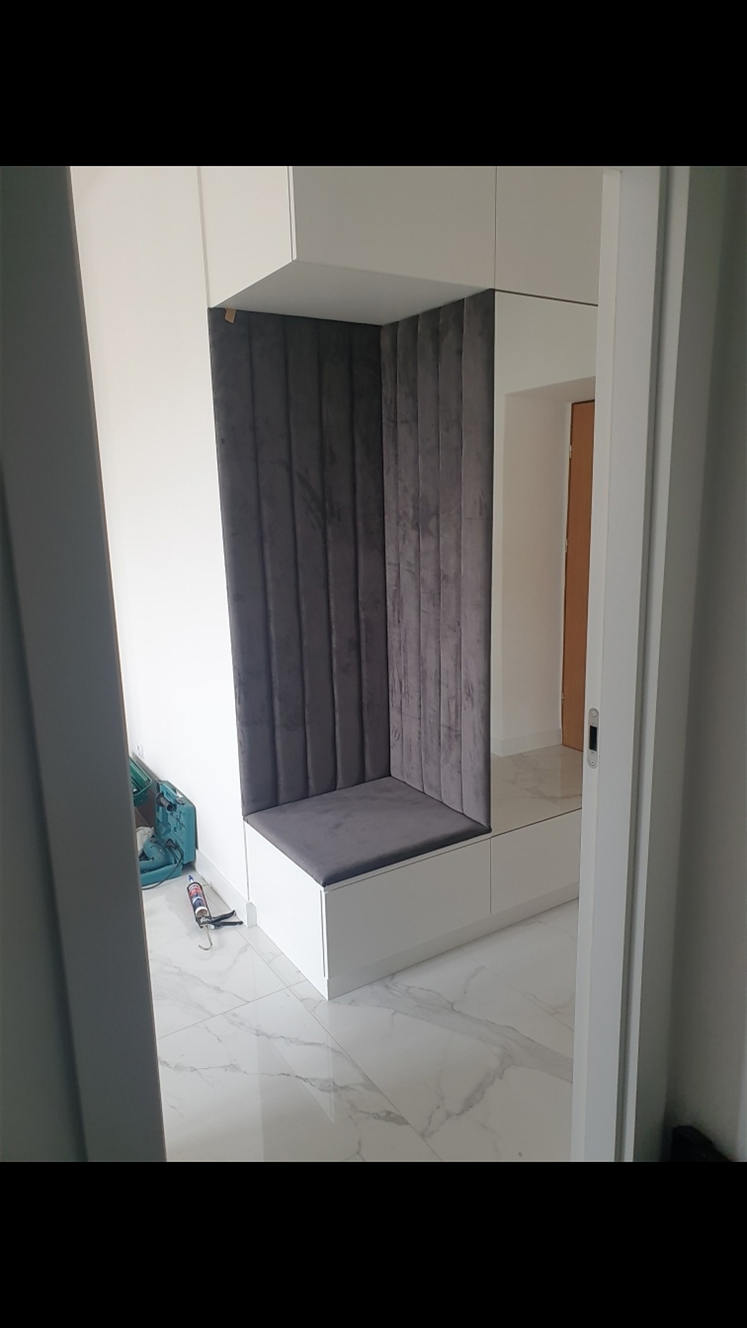 Wnęka z siedziskiem obita szarą, welurową tkaniną w pionowe pasy, wkomponowana w białą szafę w przedpokoju, widok zza futryny drzwi.