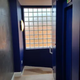 Korytarz z granatowymi ścianami, szarą ścianą z abstrakcyjnymi rysami, jasną podłogą z paneli, oświetleniem sufitowym i lampą ścienną, oraz oknem z luksferami.