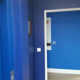 Wnętrze pomalowane na intensywny odcień niebieskiego, widoczna rama drzwiowa z białymi listwami przypodłogowymi i drewniana podłoga, z boku fragment windy z panelem sterowania.