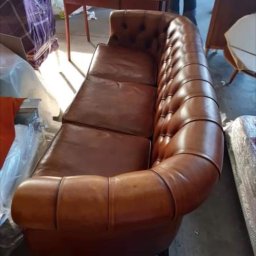 KASHICHIDOSHI Sp.z.o.o - Skórzana sofa Chesterfield w odcieniach brązu, widoczna w magazynie przed załadunkiem, z elementami mebli w tle.