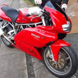 KASHICHIDOSHI Sp.z.o.o - Czerwony motocykl Ducati 1000 zaparkowany na chodniku, z kartonem w tle, prawdopodobnie przygotowany do przewozu.