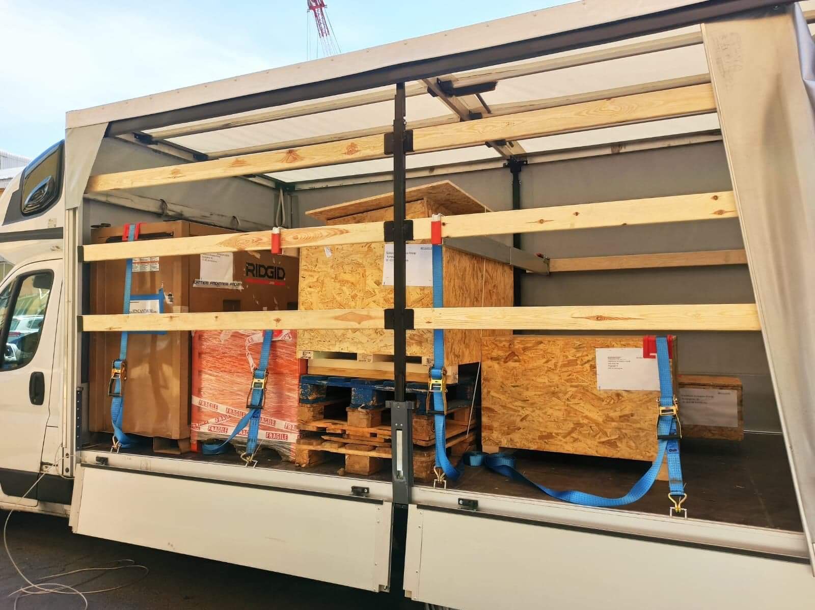 Wnętrze paki busa załadowane różnymi paczkami i skrzyniami, zabezpieczonymi niebieskimi pasami transportowymi i drewnianą konstrukcją, widoczny fragment budynku w tle.