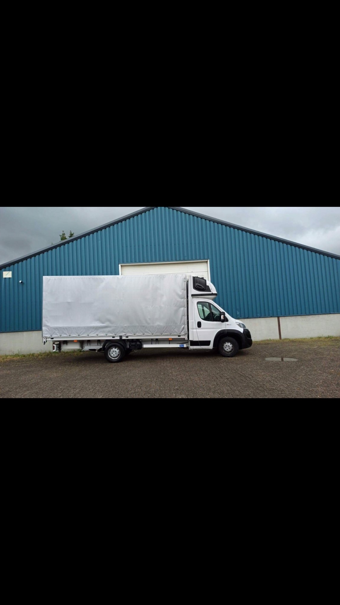 Biały samochód dostawczy z plandeką na tle niebieskiego magazynu z blachy falistej.