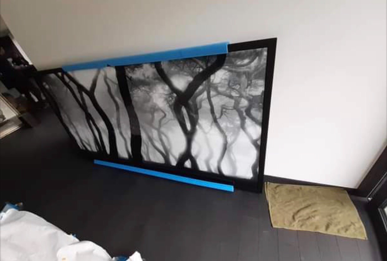 Duży obraz w czarnej ramie z motywem drzew, zabezpieczony niebieską pianką, leży na podłodze podczas przeprowadzki.