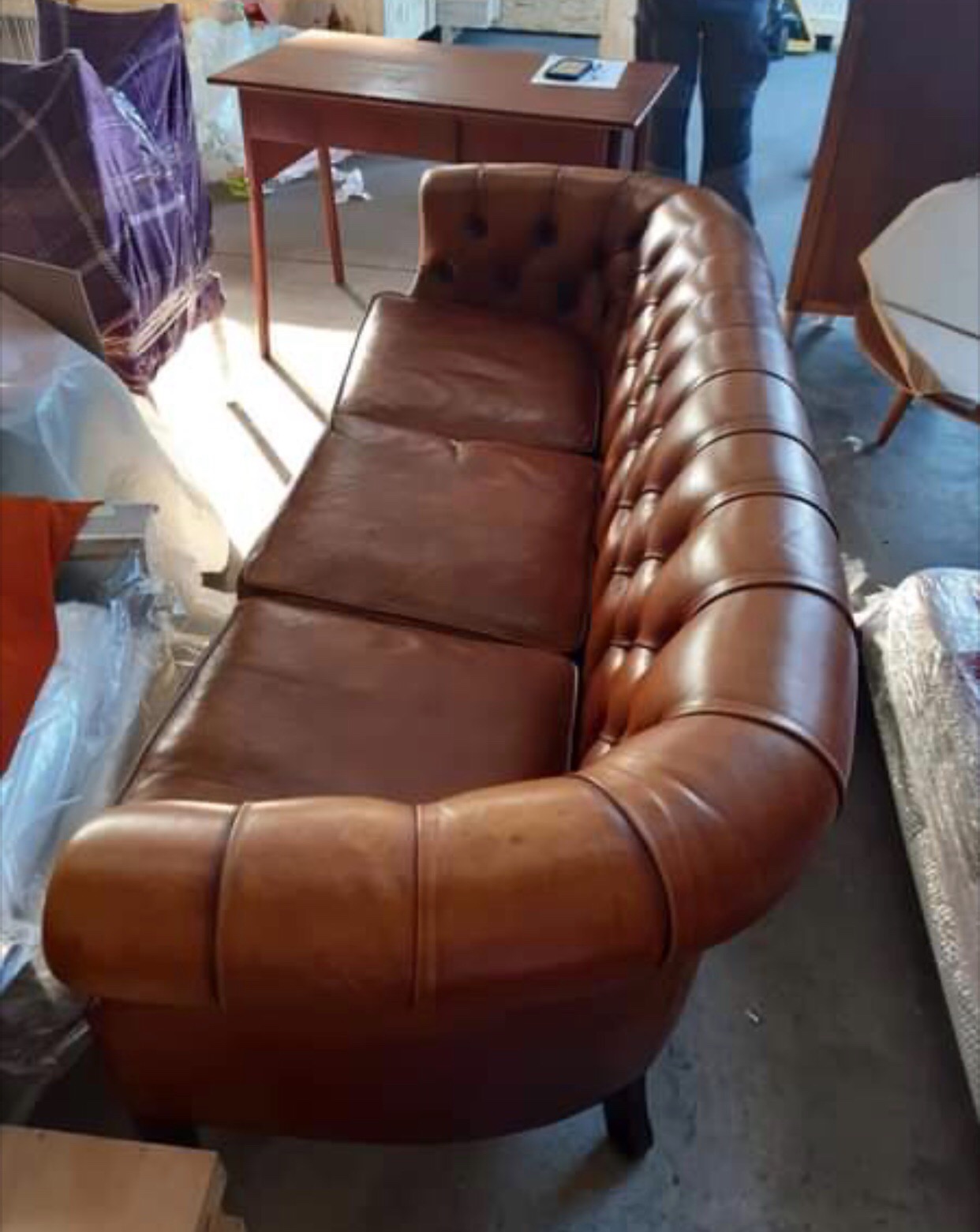 Skórzana sofa Chesterfield w odcieniach brązu, widoczna w magazynie przed załadunkiem, z elementami mebli w tle.