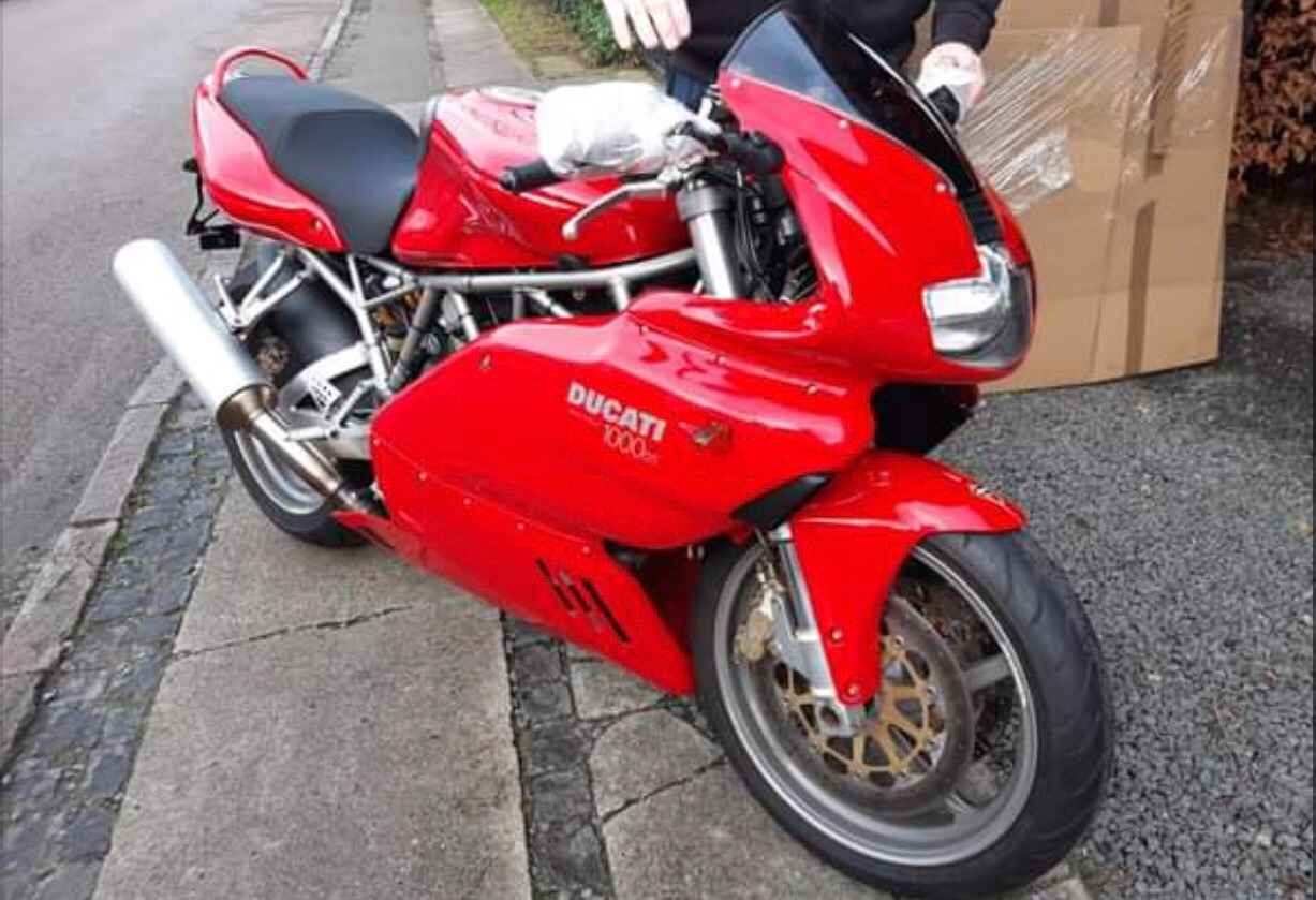 Czerwony motocykl Ducati 1000 zaparkowany na chodniku, z kartonem w tle, prawdopodobnie przygotowany do przewozu.