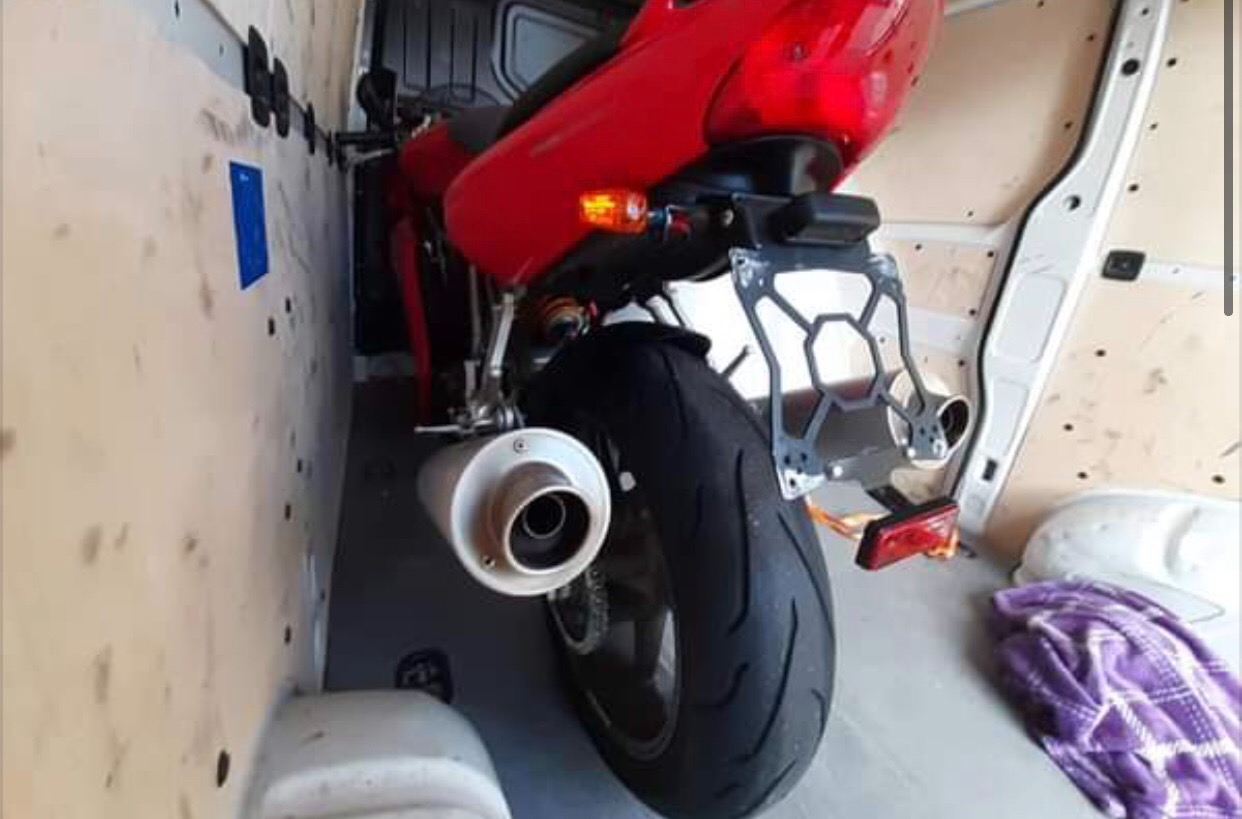 Czerwony motocykl sportowy, widok z tyłu, zabezpieczony wewnątrz furgonetki wyłożonej sklejką, z widocznym tłumikiem i tylnym światłem.