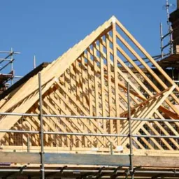 Konstrukcja drewniana dachu w trakcie budowy, widoczne elementy więźby dachowej, rusztowania i komin z cegły w tle, jasne, słoneczne oświetlenie.