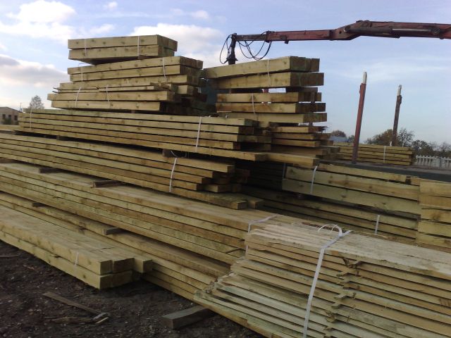 Stosy impregnowanego drewna budowlanego ułożone na placu, widoczny dźwig z hakiem w tle, zabezpieczone białymi taśmami.