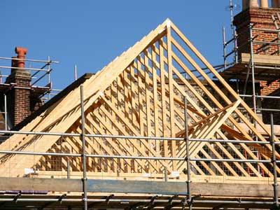 Konstrukcja drewniana dachu w trakcie budowy, widoczne elementy więźby dachowej, rusztowania i komin z cegły w tle, jasne, słoneczne oświetlenie.