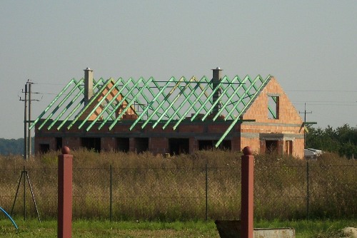 Surowy stan budowy domu jednorodzinnego z widoczną konstrukcją dachu z impregnowanego drewna w kolorze zielonym, ceglanymi ścianami i kominem oraz otaczającym terenem z wysoką trawą.