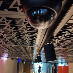 MEDIA M PATRYK KOWALCZUK - Czarna kamera kopułkowa zamontowana na suficie z ozdobnym wzorem, widoczne także reflektory i sylwetki osób w tle.