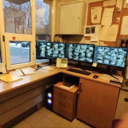 MEDIA M PATRYK KOWALCZUK - Stanowisko pracy z trzema monitorami wyświetlającymi obraz z kamer monitoringu, biurko z notatkami, okno z widokiem na zewnątrz, tablica korkowa z dokumentami i schematami na ścianie.