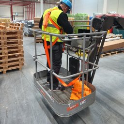 MEDIA M PATRYK KOWALCZUK - Pracownik w hełmie i kamizelce na podnośniku koszowym w hali magazynowej, prawdopodobnie podczas montażu instalacji pod sufitem.