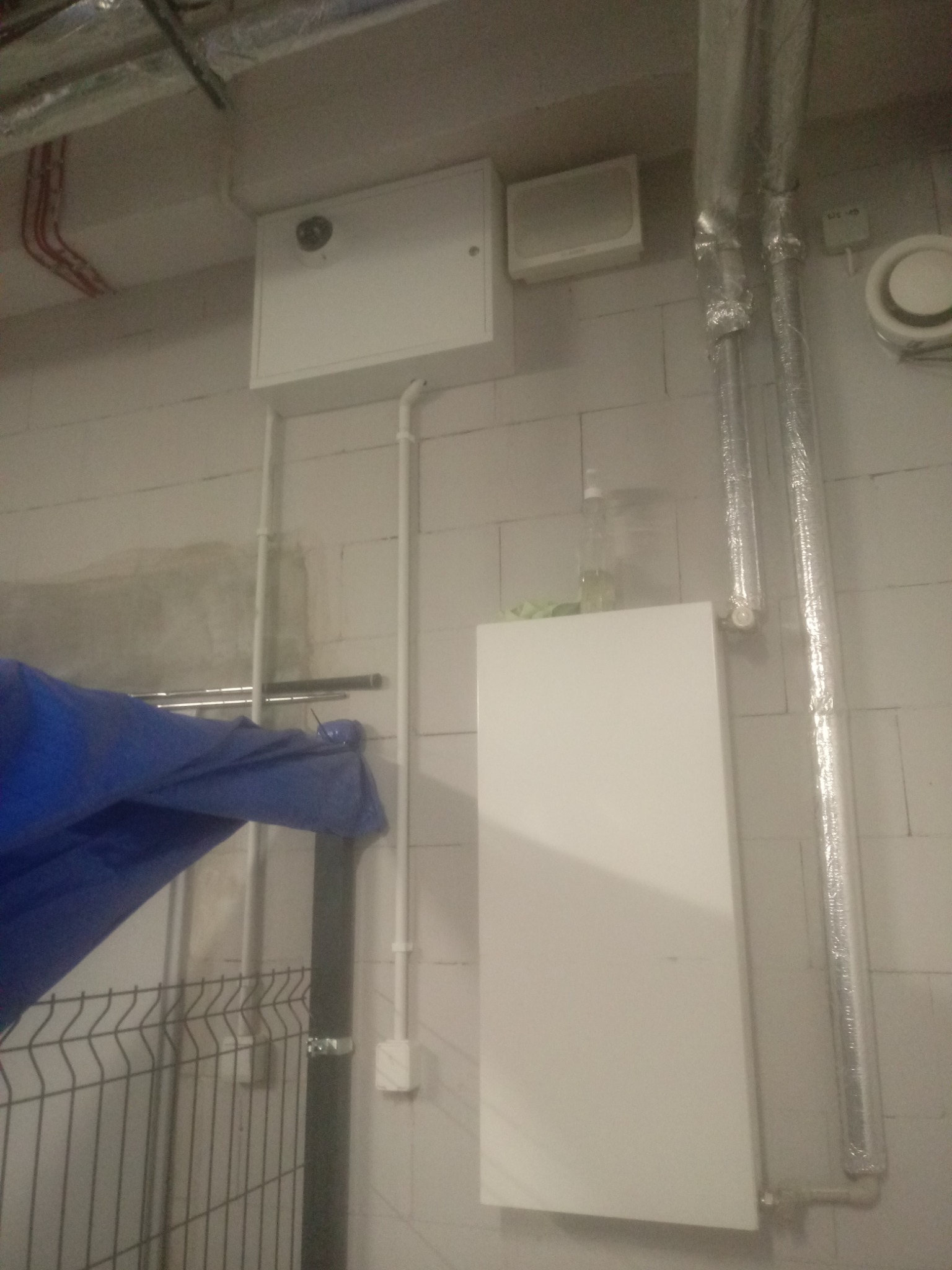 Biała skrzynka z kamerą na ścianie z bloków, obok rury w srebrnej izolacji i niebieska plandeka