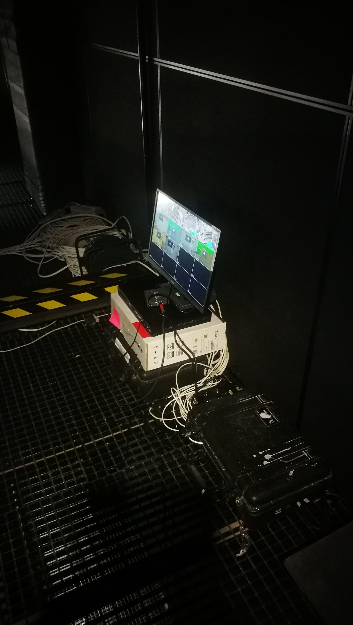 Ekran monitora wyświetlający obraz z kamer, ustawiony na kartonie i urządzeniach elektronicznych, otoczony plątaniną kabli w ciemnym pomieszczeniu z metalową kratą podłogową.
