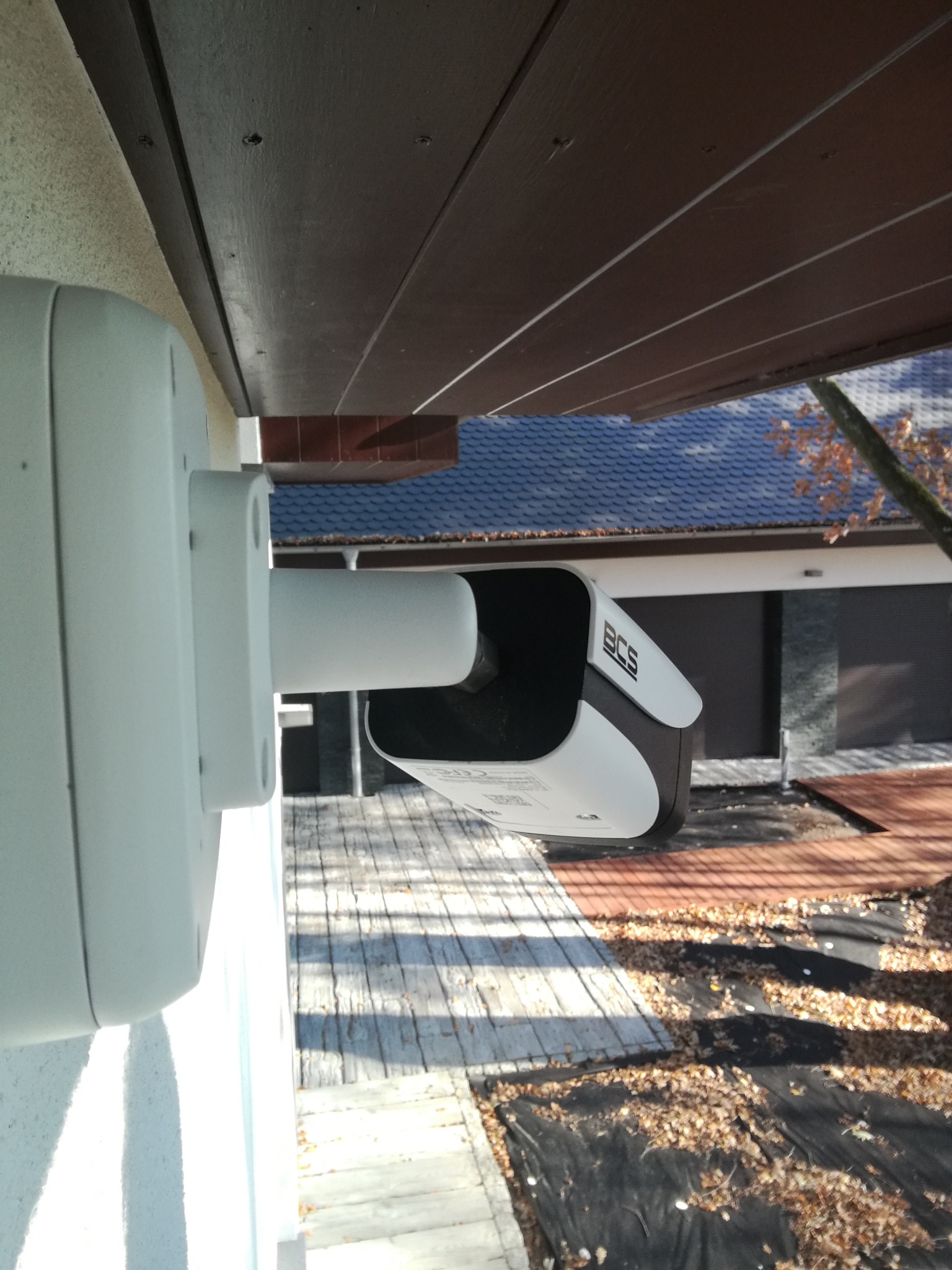 Zewnętrzna kamera monitoringu BCS zamontowana pod okapem budynku z widocznym otoczeniem posesji pokrytym opadłymi liśćmi.