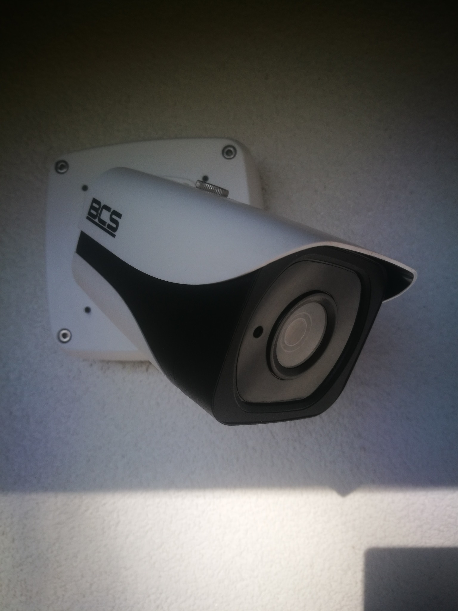 Biała kamera monitoringu BCS zamontowana na jasnej ścianie, widok z dołu.