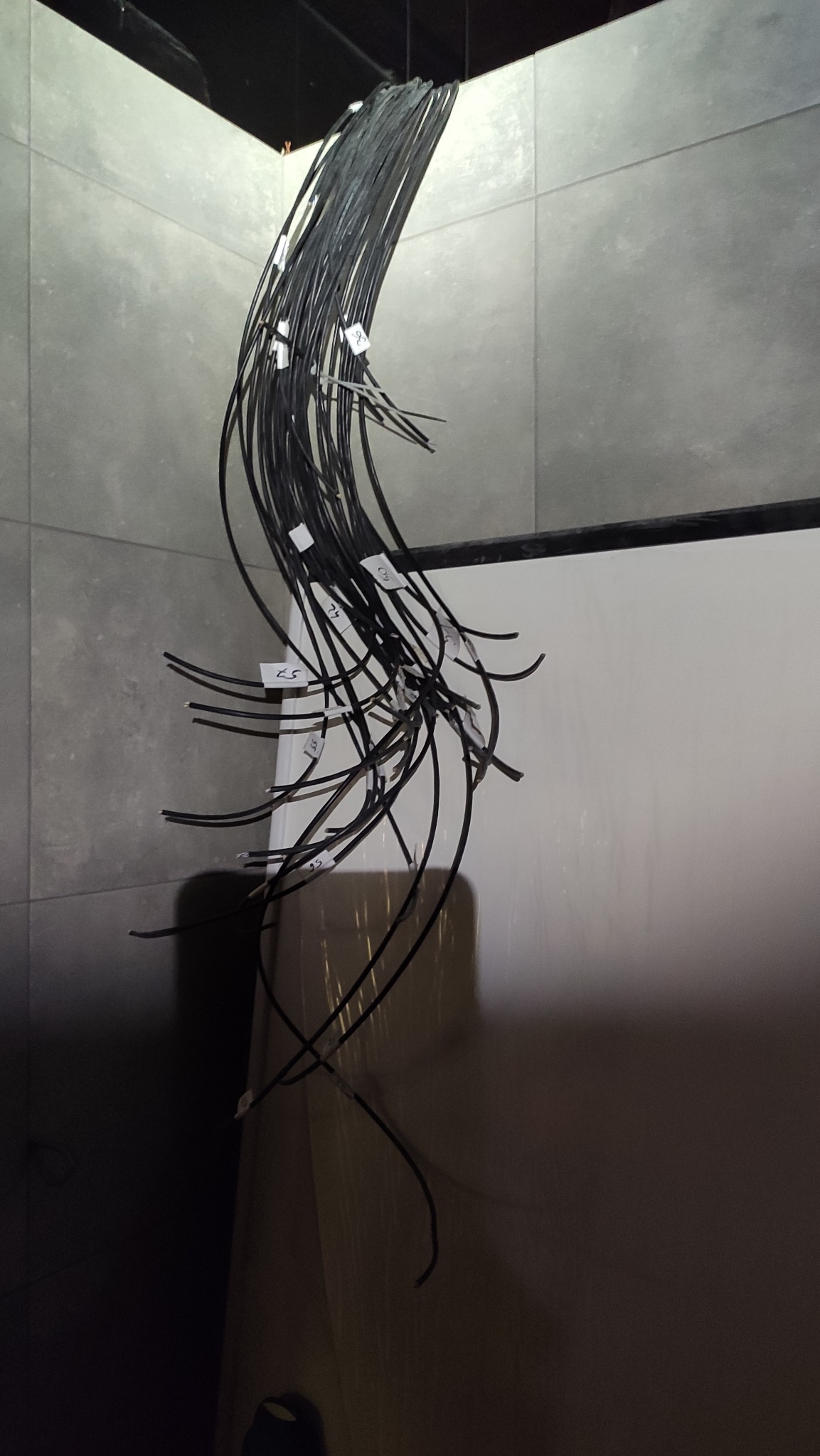 Pęk czarnych kabli zwisających z sufitu, oznaczonych białymi etykietami, na tle szarej ściany z płytek.