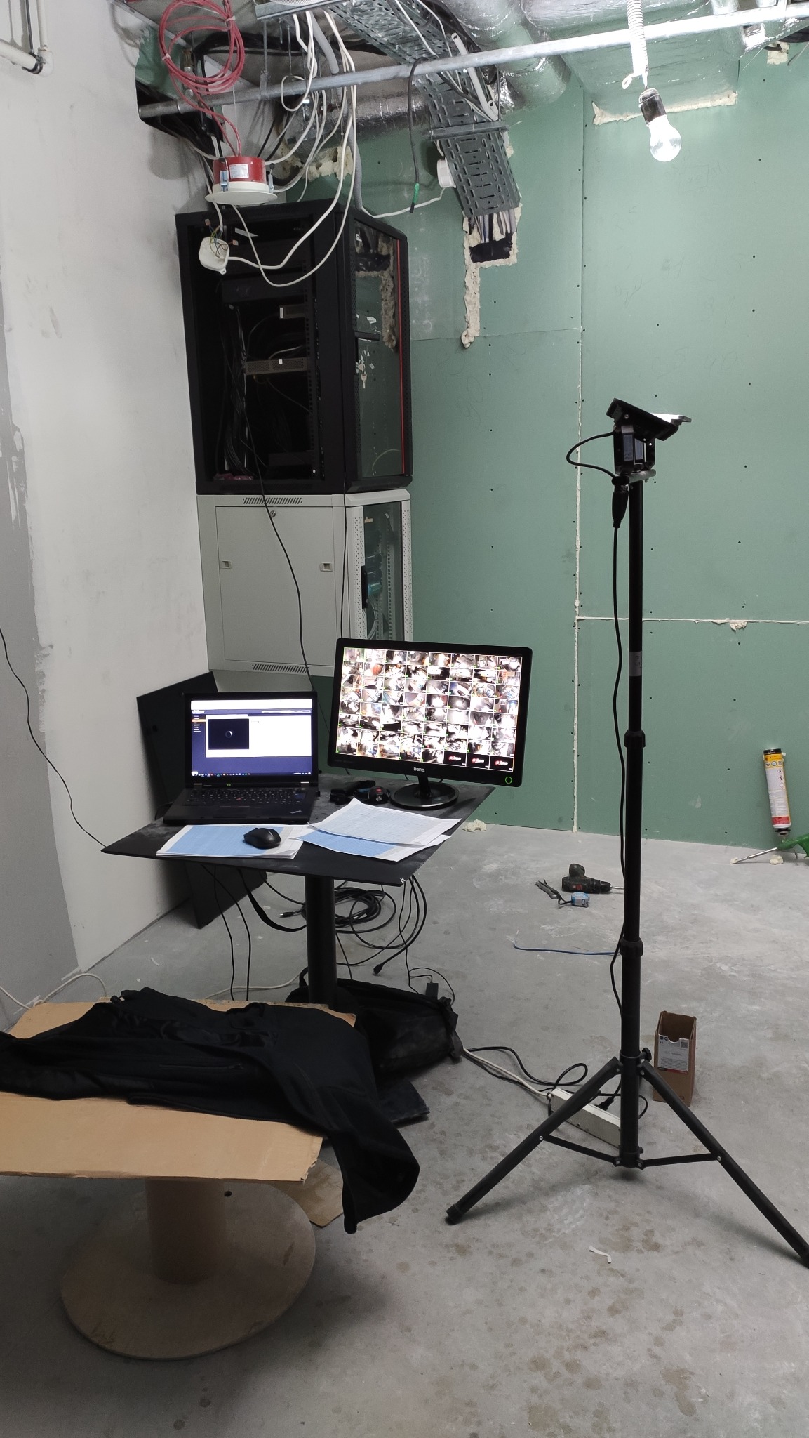 Stanowisko robocze podczas instalacji monitoringu: laptop, monitor z podglądem z kamer, kamera na statywie, serwerownia w tle, surowy stan wykończenia wnętrza.
