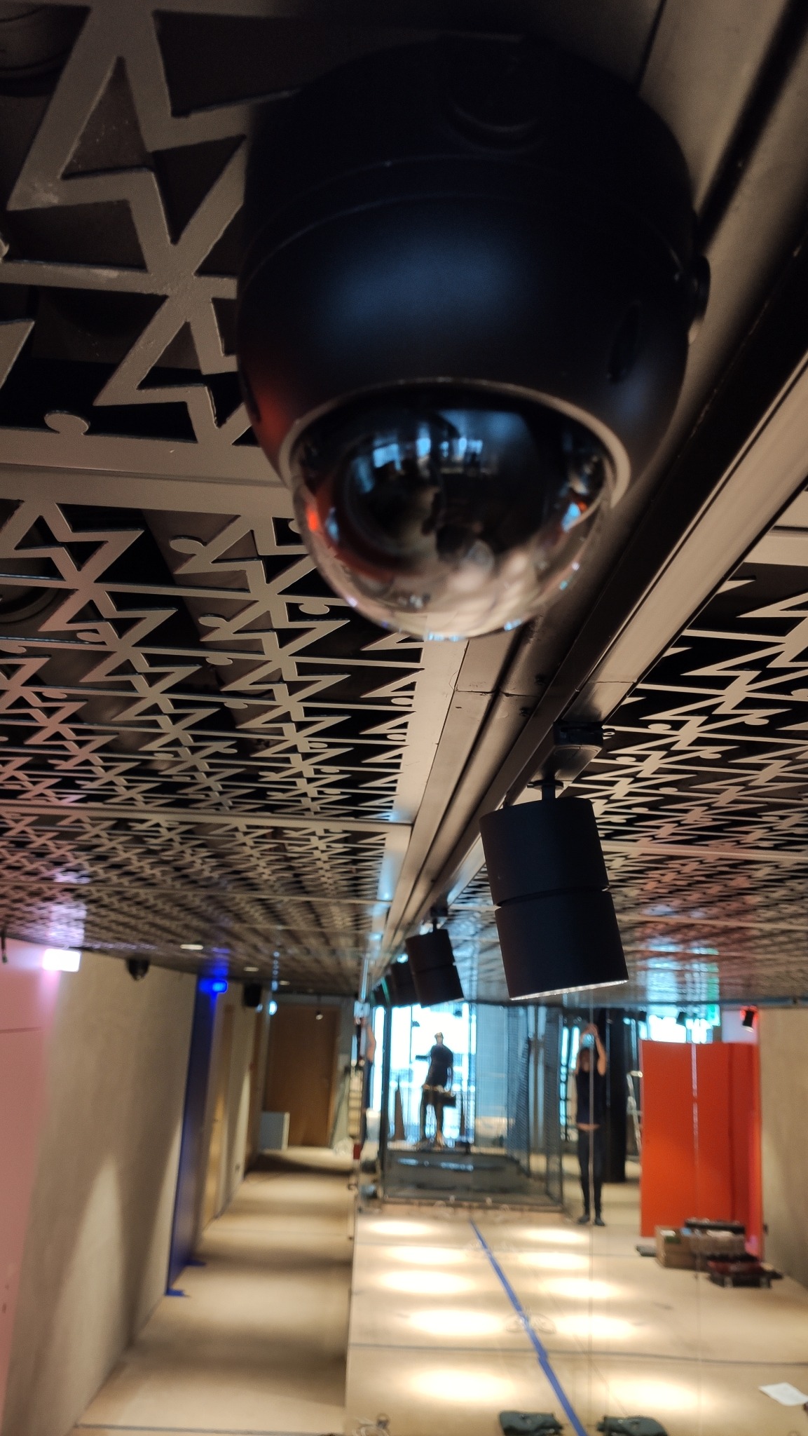 Czarna kamera kopułkowa zamontowana na suficie z ozdobnym wzorem, widoczne także reflektory i sylwetki osób w tle.