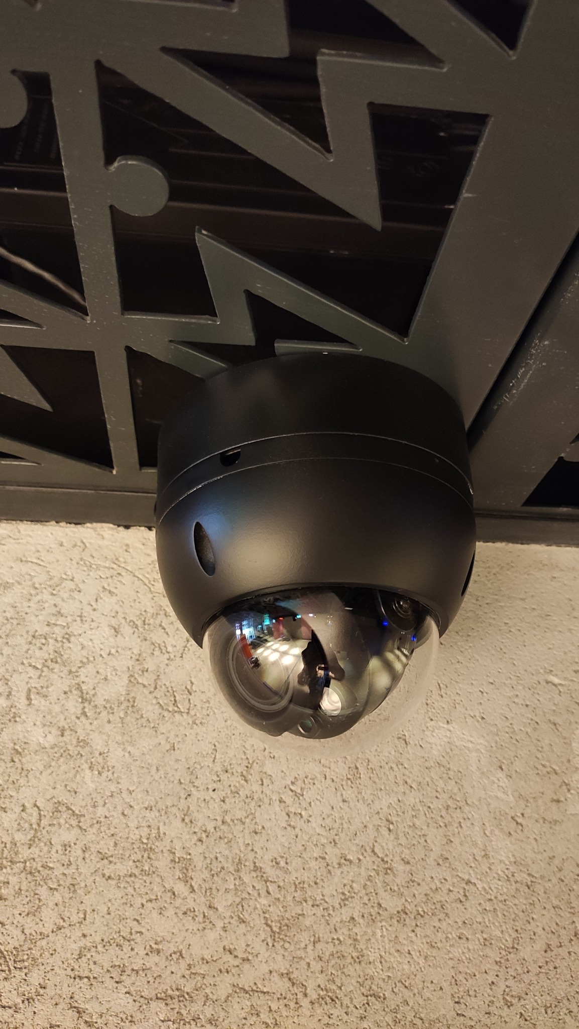 Czarna kamera kopułkowa monitoringu zamontowana pod ażurowym sufitem w Nowej Woli, widoczne odbicie otoczenia w obiektywie.