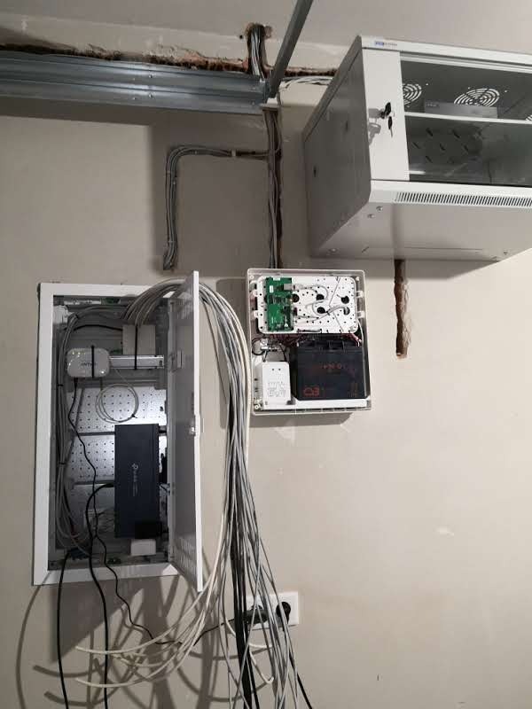 Rozdzielnia elektryczna z licznymi kablami w białej obudowie, obok skrzynka z akumulatorem i płytką drukowaną, nad nimi szafka rack z kluczem w zamku, widoczne kable prowadzone po ścianie i suficie.