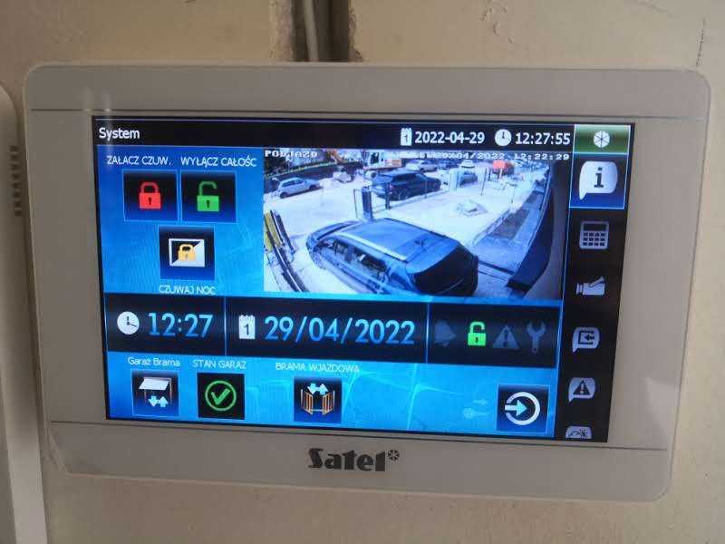 Ekran dotykowy systemu alarmowego Satel z podglądem z kamery monitoringu parkingu, wyświetlający datę i godzinę 29.04.2022 12:27, oraz statusy alarmu i bramy garażowej.