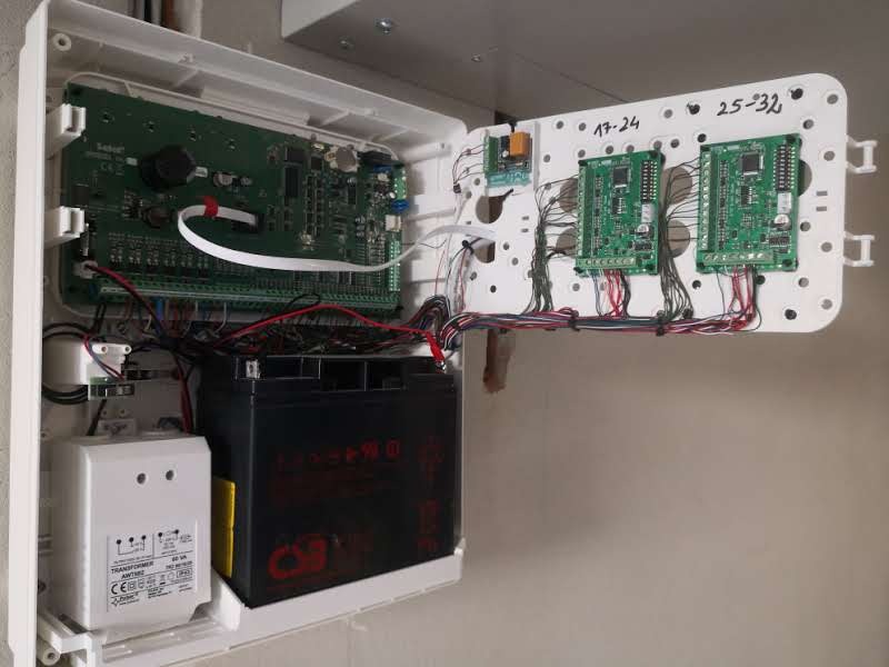 Otwarta obudowa centrali alarmowej z widoczną elektroniką, akumulatorem i podłączonymi przewodami, w trakcie instalacji.