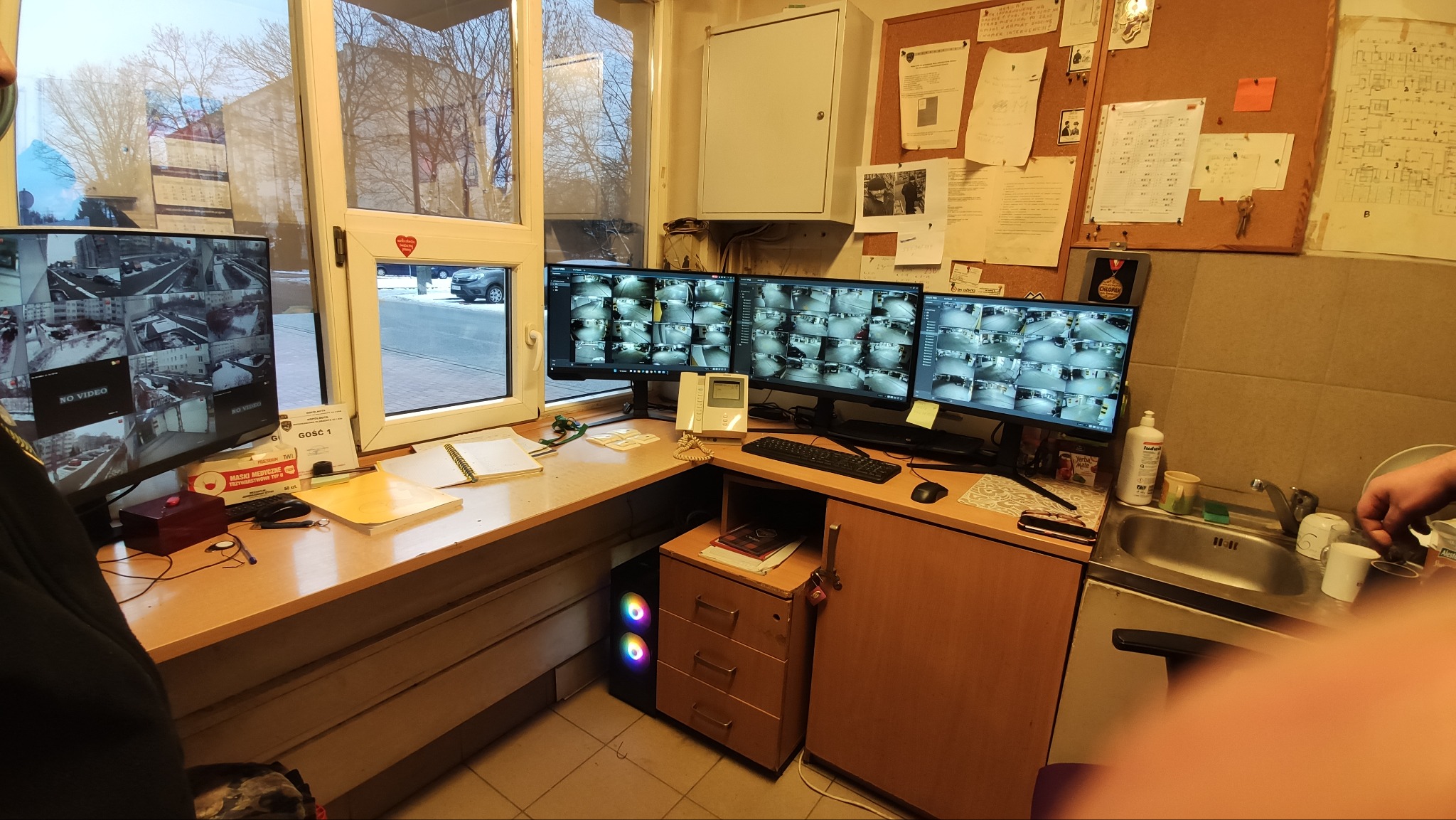 Stanowisko pracy z trzema monitorami wyświetlającymi obraz z kamer monitoringu, biurko z notatkami, okno z widokiem na zewnątrz, tablica korkowa z dokumentami i schematami na ścianie.