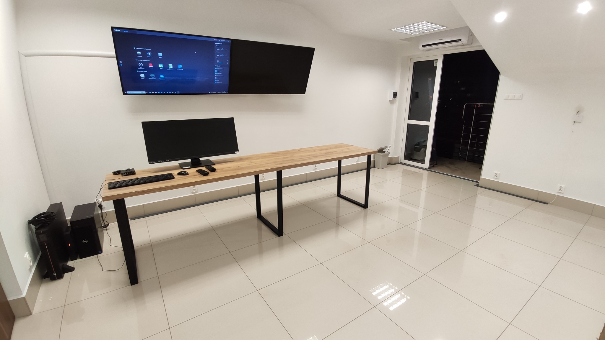 Sala konferencyjna z dwoma monitorami na ścianie, biurkiem, komputerem stacjonarnym i widokiem na zewnątrz przez przesuwne drzwi.