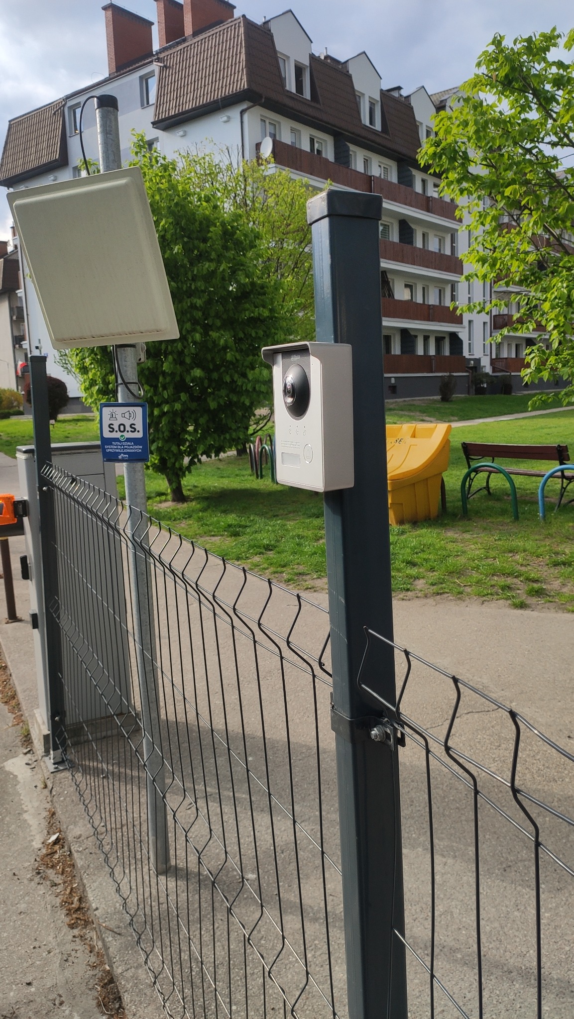 Nowoczesny wideofon zamontowany na słupku ogrodzenia panelowego, na tle budynku mieszkalnego i zieleni.