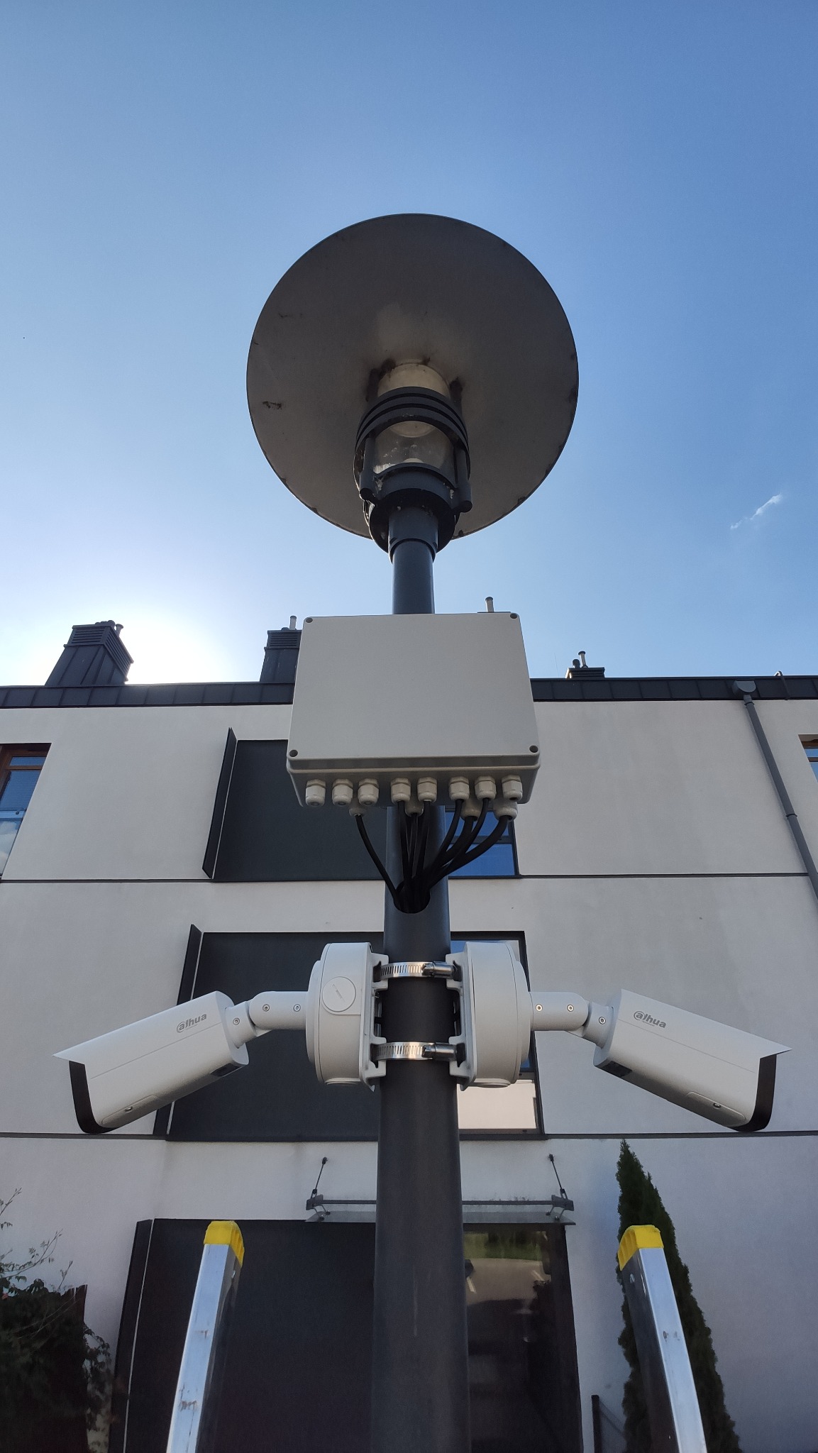 Dwie kamery monitoringu Dahua zamontowane na słupie oświetleniowym z puszką przyłączeniową, na tle budynku z białą elewacją i fragmentem drabiny.