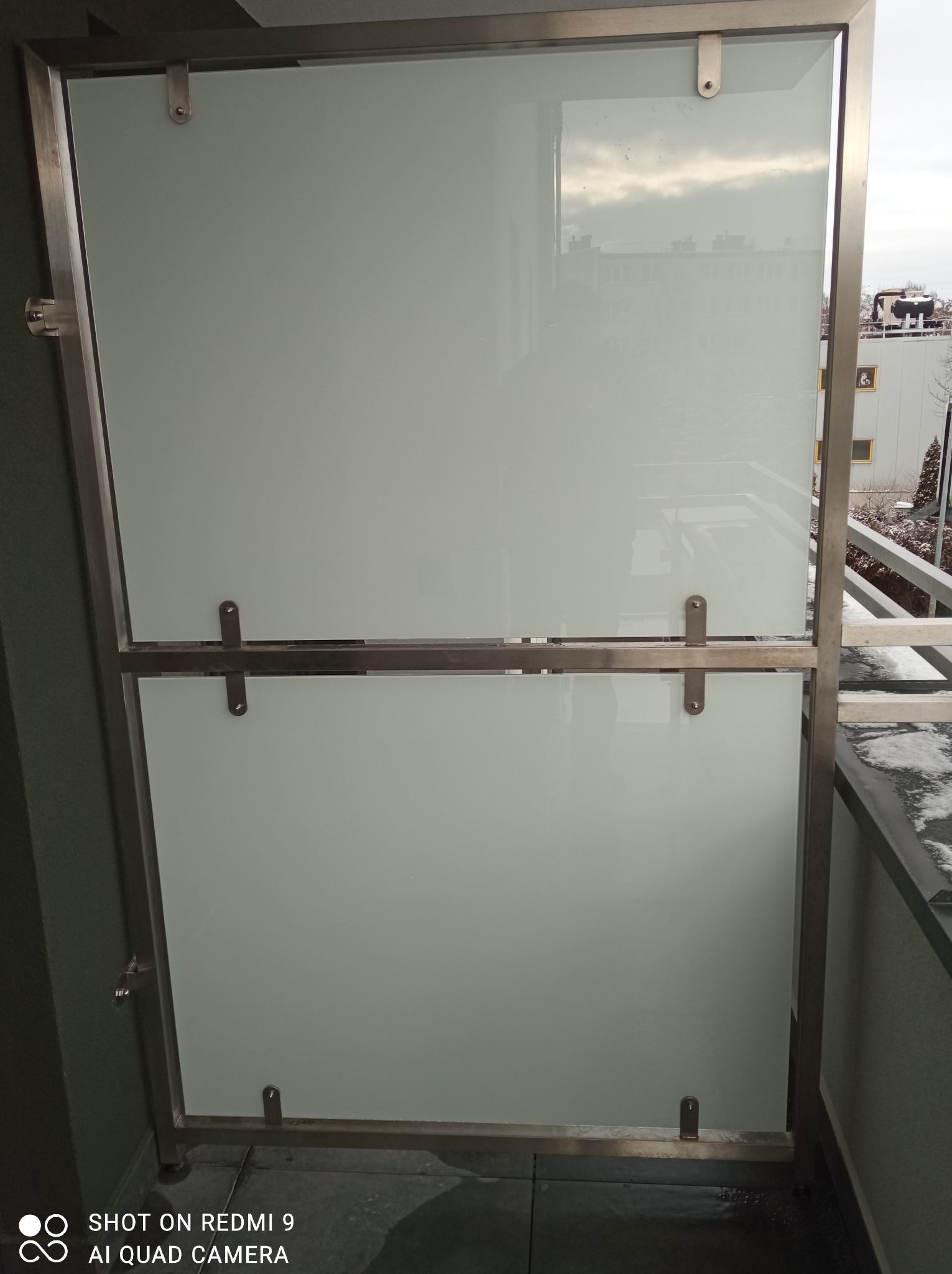 Szklana osłona balkonowa z nierdzewną ramą, zamontowana na balkonie z widokiem na budynki i zimowy krajobraz.