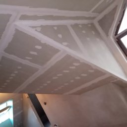 Wykończenia wnętrz. - Sufit poddasza wykończony płytami gipsowo-kartonowymi, widoczne łączenia szpachlowane, przygotowane do dalszych prac wykończeniowych, okno dachowe w prawym górnym rogu.