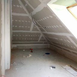 Wykończenia wnętrz. - Poddasze w trakcie wykańczania: zamontowane płyty gipsowo-kartonowe z widocznymi łączeniami przed szpachlowaniem, przewody elektryczne zwisające ze ściany, narzędzia pracy (wiertarka) i materiały...