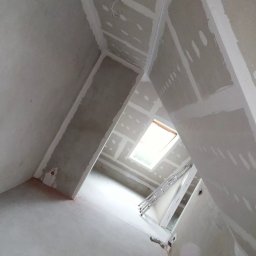Wykończenia wnętrz. - Wnętrze poddasza w trakcie wykańczania, widoczne płyty gipsowo-kartonowe na skośnych ścianach i suficie, szpachlowane łączenia, okno dachowe i rozstawiona aluminiowa drabina.