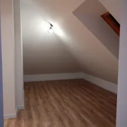 Poddasze ze skośnym sufitem, wykończone panelami podłogowymi w odcieniach brązu i beżu, oświetlone pojedynczą, nowoczesną lampą wiszącą. Widoczne okno dachowe w drewnianej ramie.