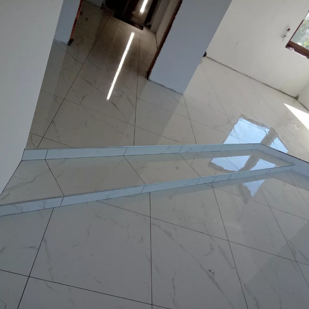 Ułożone, błyszczące, białe płytki imitujące marmur na podłodze z widocznymi, srebrnymi listwami progowymi i odbiciem okna w powierzchni.