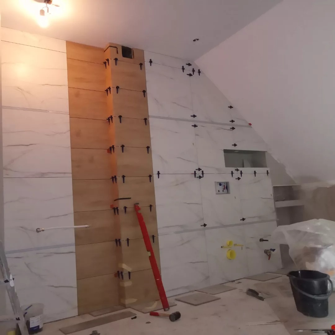 Montaż płytek imitujących marmur i drewno w łazience na poddaszu, widoczne krzyżyki dystansowe, poziomica i młotek gumowy.