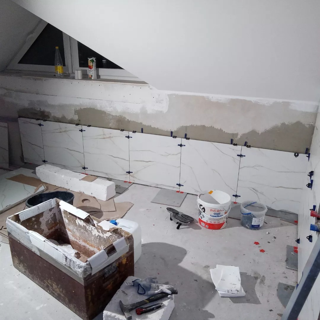 Montaż białych płytek imitujących marmur na ścianie w łazience na poddaszu, widoczne krzyżyki dystansowe, narzędzia glazurnicze i wiaderka z klejem.