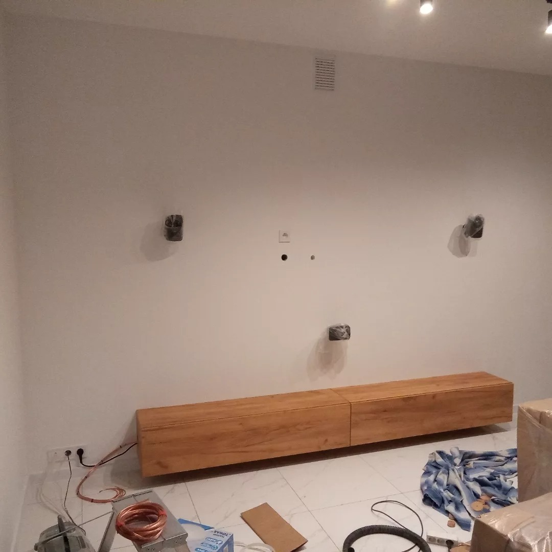 Montaż wiszącej szafki RTV z drewnianym fornirem na białej ścianie, widoczne przygotowane punkty elektryczne, przewody, narzędzia i materiały na marmurowej podłodze.