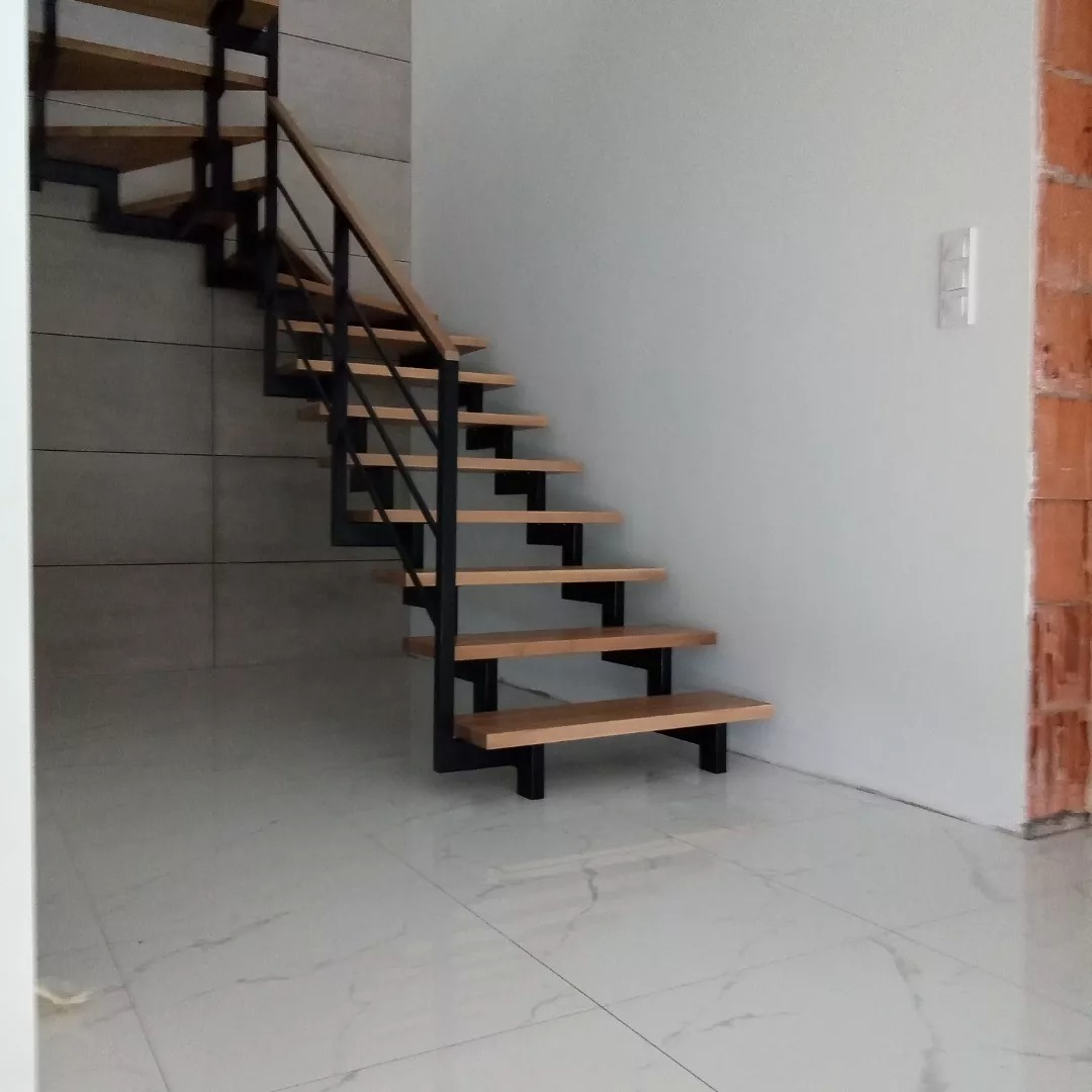 Nowoczesne schody wewnętrzne z drewnianymi stopniami i czarną metalową balustradą, widoczne na tle białej ściany z włącznikiem światła i fragmentu ściany z czerwonej cegły.