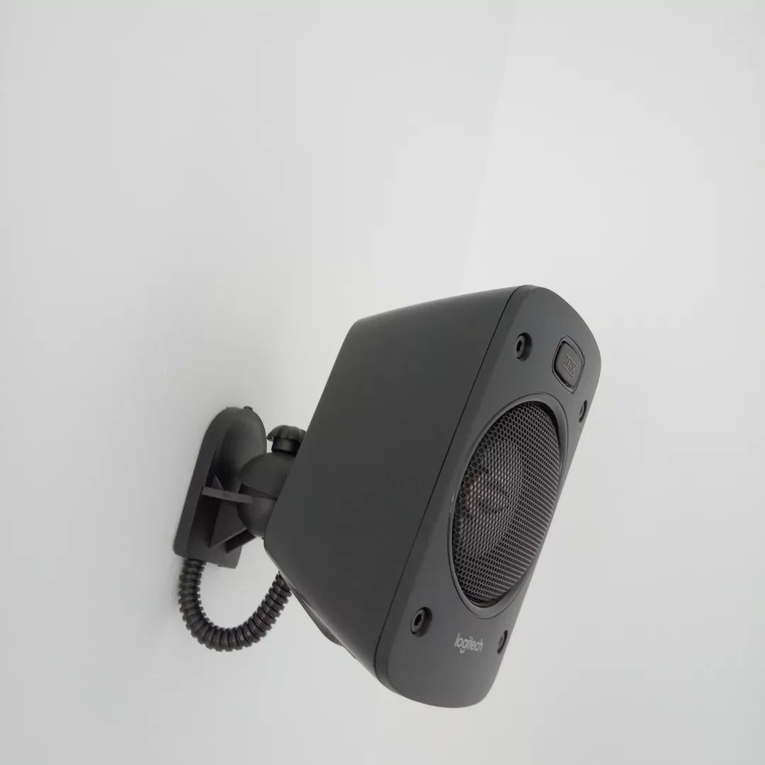 Czarny głośnik marki Logitech zamontowany na ścianie za pomocą uchwytu z kablem w osłonie karbowanej.