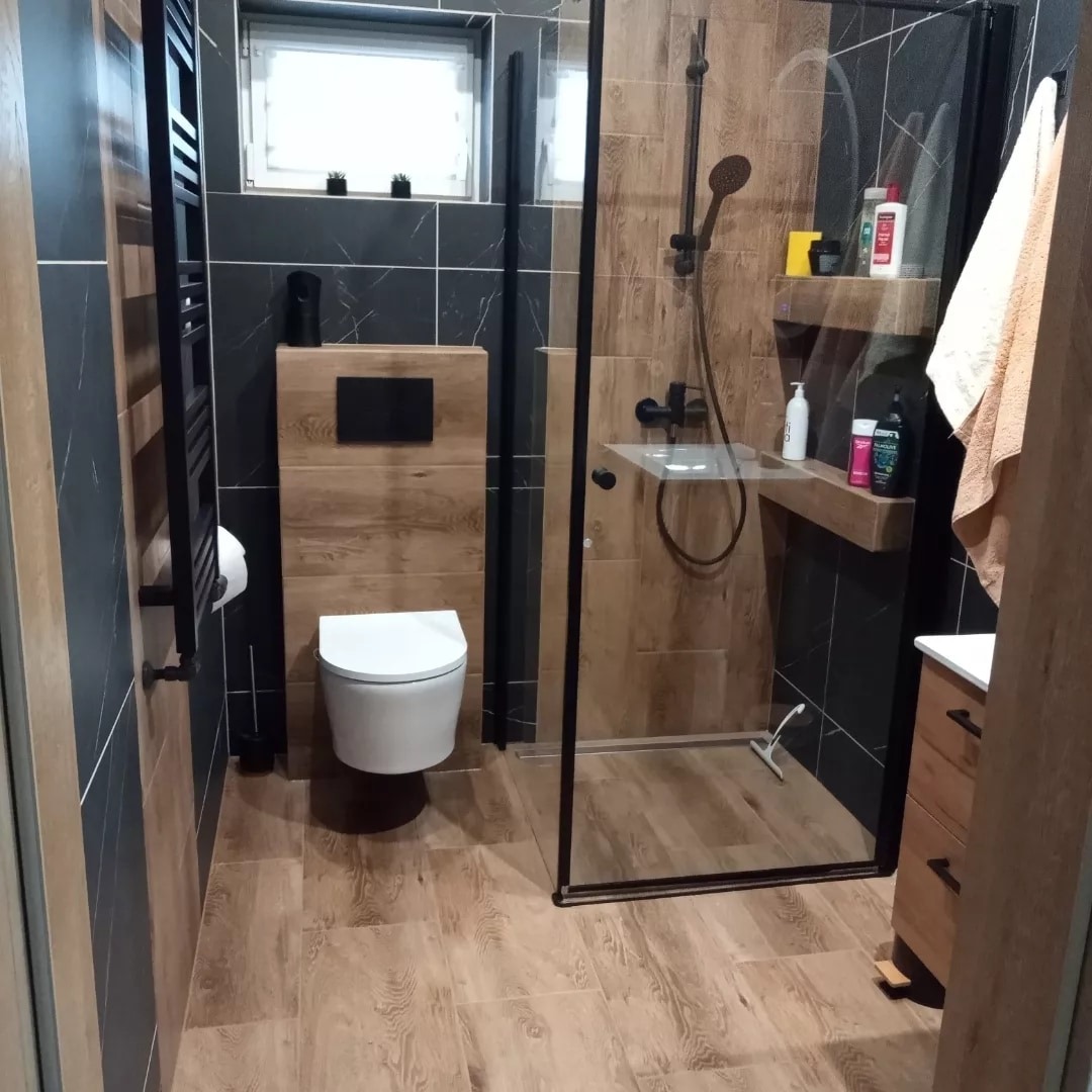 Nowoczesna łazienka z czarnymi i drewnopodobnymi płytkami, kabiną prysznicową typu walk-in z czarną ramą, podwieszaną toaletą i grzejnikiem drabinkowym w kolorze czarnym.