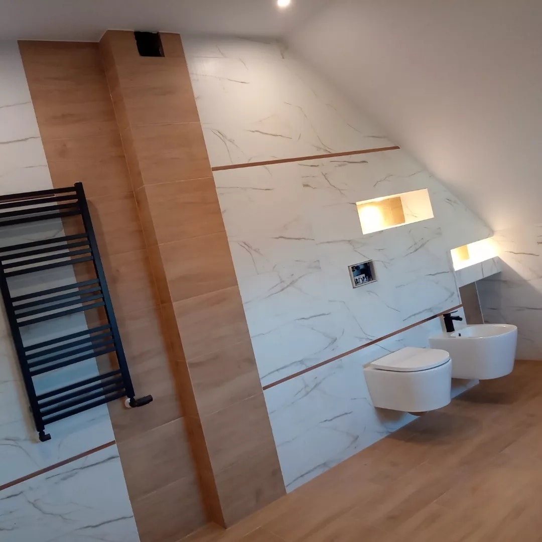 Nowoczesna łazienka na poddaszu z biało-marmurowymi płytkami, drewnopodobną podłogą, podwieszaną toaletą i bidetem oraz czarnym grzejnikiem drabinkowym.