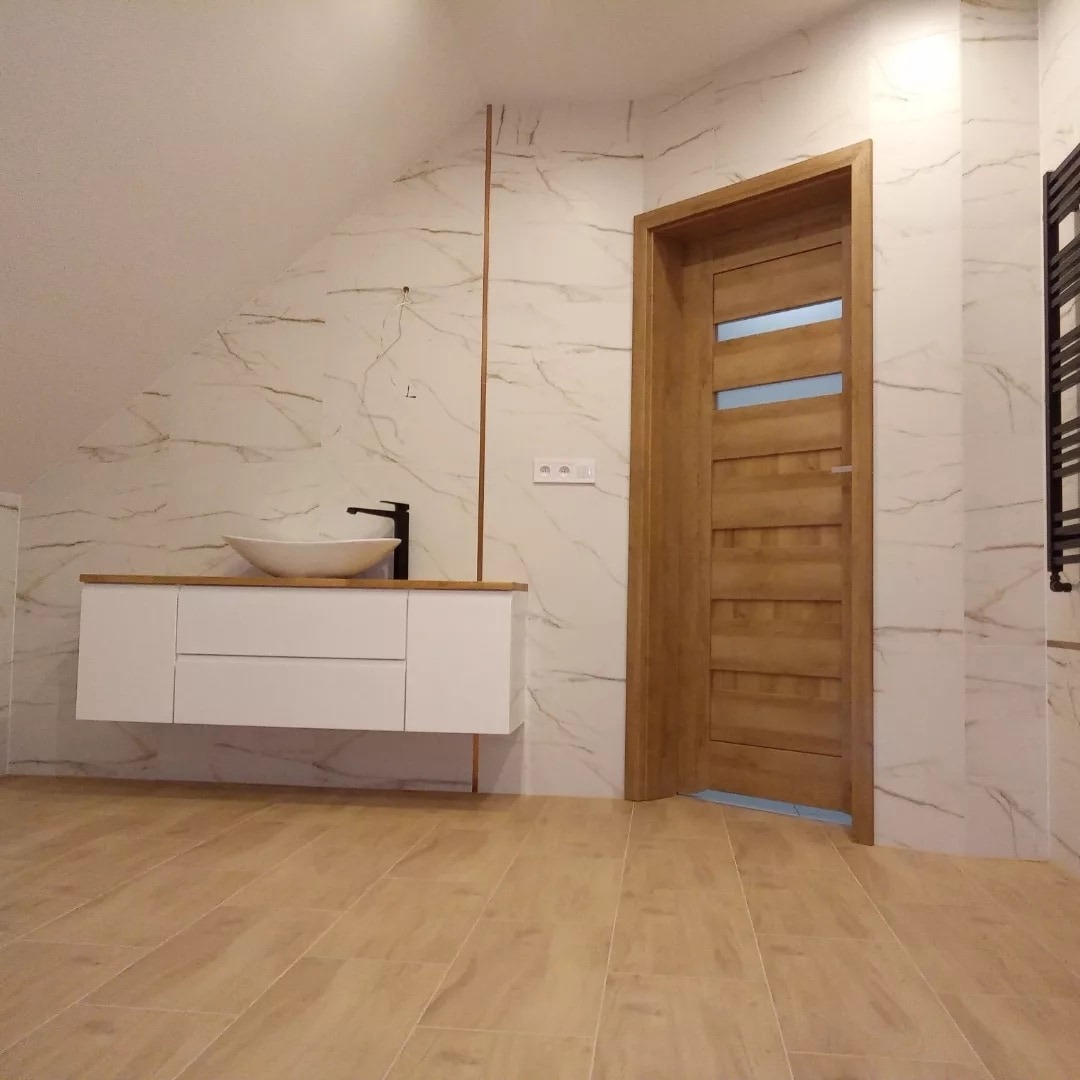 Nowoczesna łazienka na poddaszu z umywalką nablatową, białymi szafkami pod umywalką z drewnianym blatem, marmurowymi płytkami na ścianach, drewnianymi drzwiami z przeszkleniami i podłogą z płytek...