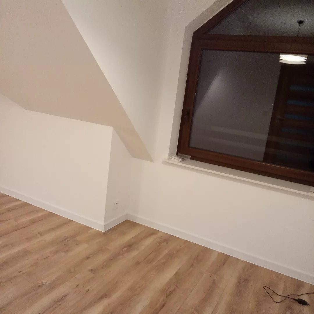 Poddasze po remoncie: Ściany pomalowane na biało, drewniana podłoga, okno dachowe z brązową ramą. Widoczna listwa przypodłogowa i gniazdko elektryczne.